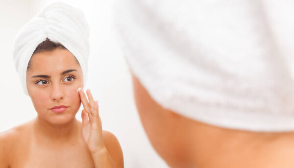 Portrait of a beautiful young woman applying face cream and looking at mirror in bathroom. Youthful girl with perfect skin using facial cosmetics. Skin care and health concept
