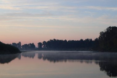 Güzel, sakin bir gün doğumu. Nehir kenarında, ayna suyunun yansımasıyla. Pastel renkler, doğa manzarası. Ağaçlar siluet, sis, bulutlar ve günbatımı fotoğrafı.