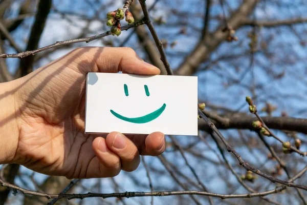 Happy green smile emoji on white paper card holding in hand on sunny spring blue sky background. Positive mood good weather time