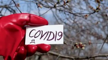 Covid19 Karantina beyaz kart değişiminde evde kalın. Eller kırmızı koruyucu eldivenlerle güneşli bahar ağaçlarının arka planında. Temiz hava tehlikeyi önler, tecrit bilgilerini koru