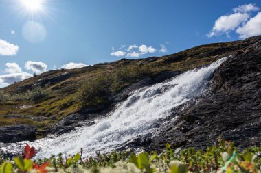 Güneşli beyaz manzaralı Norveç güçlü nehir şelalesi koyu kayalar, parlak mavi gökyüzü ve bulanık çimenler