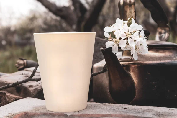 White plastic cup glass with aroma flavored tea and old grunge soot vintage teapot. Spring cherry blossom white flowers. Lifestyle nature outdoors recreation picnic mood. Color graded