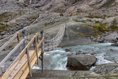 Dağ nehri üzerindeki tahta köprü. Jostedalsbreen Ulusal Parkı 'ndaki Nigardsbreen' e bulutlu bir gezi. Norveç dağları mavi su nehri, kayalar