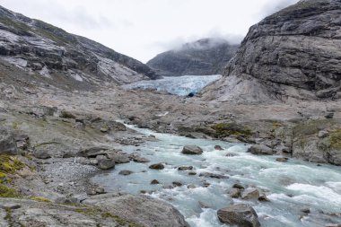 Jostedalsbreen Ulusal Parkı 'ndaki Nigardsbreen' e bulutlu bir gezi. Norveç dağlarının manzarası mavi su nehri, buz, kayalar, buzullar, kar. İklim değişikliği, eriyen buzullar