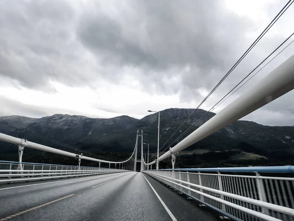 Dağlar ve fiyort yakınlarındaki gri bulutlu bir günde Norveç asfalt yolunda metal beyaz köprüden geç. Arabayla İskandinavya seyahati