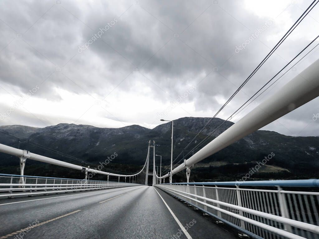 Drive Norway asphalt road across metal white bridge on grey cloudy day near mountains and fjord. Travel scandinavia by car