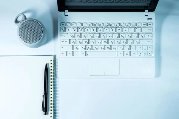 White laptop, round wireless speaker, notepad with spring binding and pen on a white background. View from above