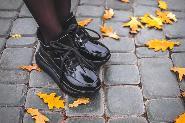 A girl stands in black shiny warm shoes on a tile with leaves in the city center in a park