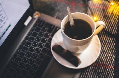 Bir fincan sıcak siyah çay, dizüstü bilgisayarında çikolata, Noel ışıkları, kış atmosferi ve rahat bir battaniye.