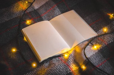 Kutsal bir kış gecesinde Noel ışıkları ve kavramsal bir fotoğrafın olduğu rahat bir ekose üzerine boş bir defter.