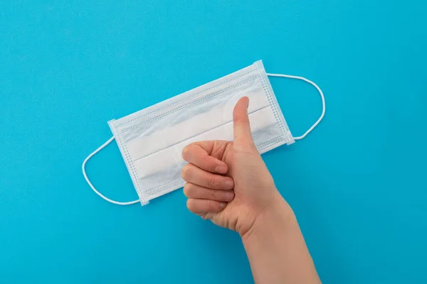 Top view of a single medical surgical mask. The doctor gives a thumbs up, approves of wearing a disposable respirator. Isolated on a blue background