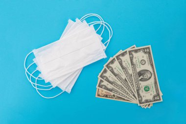 Birkaç tıbbi maskenin ve bir sürü doların üst görüntüsü. Doktorun tek kullanımlık solunum cihazı. Virüse karşı korunma maliyetini arttırma kavramı. Mavi bir arkaplanda izole edilmiş