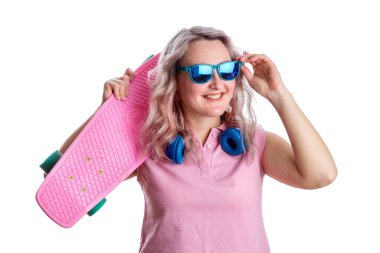 Pembe tişörtlü, kıvırcık saçlı sarışın. Pembe kaykaylı, mavi kulaklıklı ve güneş gözlüklü mutlu bir genç bayan. Beyaz bir arkaplanda izole edilmiş