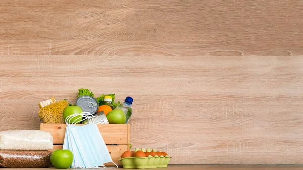 Charity donation wooden box with food with oil, milk and water, herbs, apples and oranges, cereals and canned food, rice and pasta, medical masks. Wooden background in the kitchen. Copy space for text