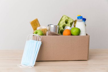 İçinde yağ, süt ve su, otlar, elmalar ve portakallar, tahıllar ve konserve yiyecekler, pirinç ve makarna ve tıbbi maskeler olan kartondan yapılmış bir bağış kutusu. Beyaz arka plan, mutfakta masa.