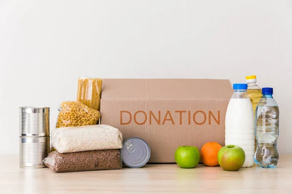 Charity donation box made of cardboard with food with oil, milk and water, herbs, apples and oranges, cereals and canned food, rice and pasta. White background, table in the kitchen