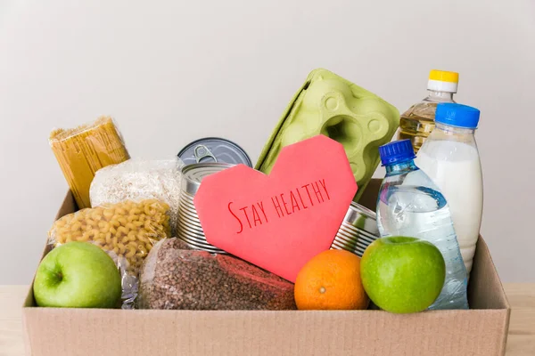 Charity donation box made of cardboard with food: oil, fruits, cereals, canned food. White background, table in the kitchen. Pink heart postcard stay healthy