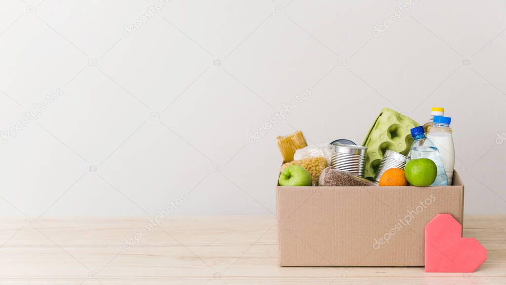 Charity donation box made of cardboard with food: oil, fruits, cereals, canned food. White background, table in the kitchen. Pink heart postcard. Copy space for text
