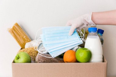 Tıbbi eldivendeki eller bağış kutusunu yiyecek ile çevirir: yağ, su, şifalı bitkiler, elma ve meyve, tahıl ve konserve yiyecek, pirinç, makarna, tıbbi maskeler. Beyaz ahşap mutfak arka planı. Kapat.