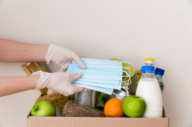 Tıbbi eldivendeki eller bağış kutusunu yiyecek ile çevirir: yağ, su, şifalı bitkiler, elma ve meyve, tahıl ve konserve yiyecek, pirinç, makarna, tıbbi maskeler. Beyaz ahşap mutfak arka planı. Kapat.