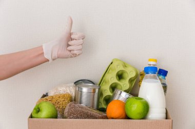 Tıbbi eldivenlerdeki eller, içinde yiyecek olan sınıf bağış kutusunu gösteriyor: yağ, su, otlar, elmalar ve meyveler, tahıllar ve konserve yiyecekler, pirinç, makarna. Beyaz ahşap mutfak arka planı. Kapat.