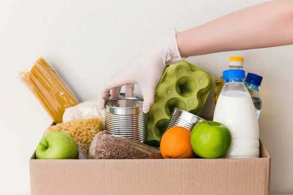 Tıbbi eldivendeki eller, içinde yiyecek, yağ, su, şifalı ot, elma ve meyve, mısır gevreği ve konserve yiyecek, pirinç ve makarna olan bağış kutusunu çeviriyor. Beyaz ahşap mutfak arka planı. Kapat.