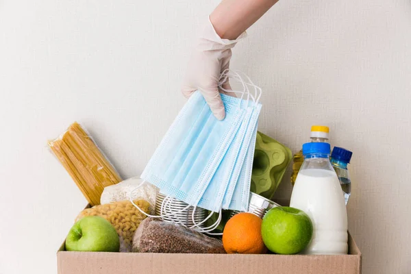 Tıbbi eldivendeki eller bağış kutusunu yiyecek ile çevirir: yağ, su, şifalı bitkiler, elma ve meyve, tahıl ve konserve yiyecek, pirinç, makarna, tıbbi maskeler. Beyaz ahşap mutfak arka planı. Kapat.