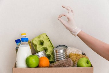 Tıbbi eldivenlerdeki eller, içinde yiyecek, yağ, su, şifalı ot, elma ve meyve, mısır gevreği ve konserve yiyecek, pirinç ve makarna olan bağış kutusu gösteriyor. Beyaz ahşap mutfak arka planı. Kapat.
