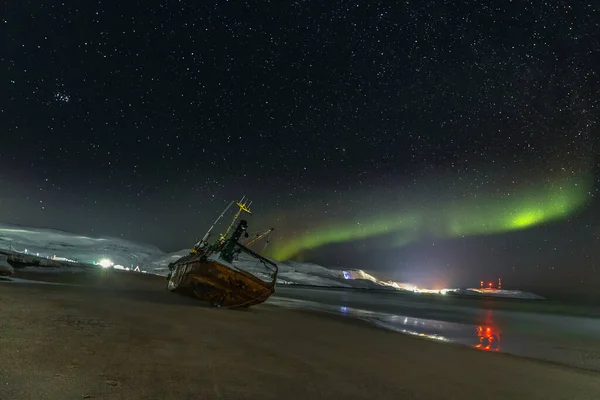 Polar lights in the starry sky. A fishing boat lying on its side, washed up by a storm on the Bank of the Barents sea. Teriberka, Russia