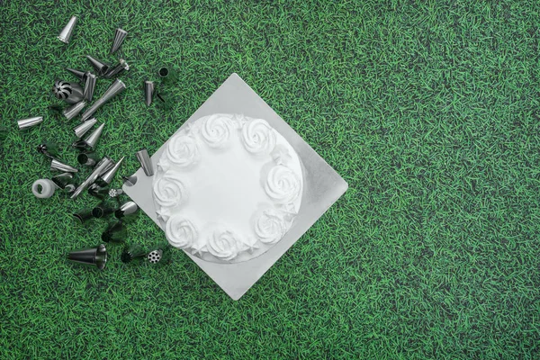 White fresh cream cake frosting with baking accessories piping nozzles flat lay on green grass background