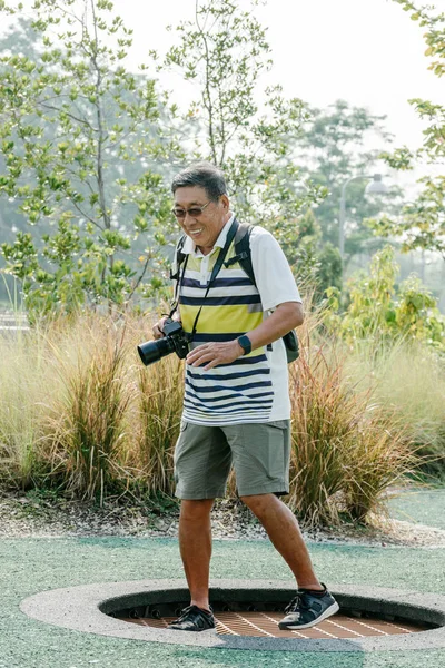 Asiatischer reifer chinesischer Rüde mit digitaler DSLR-Kamera, der sein Rentnerleben im Outdoor-Park genießt. er springt auf einem Trampolin. — Stockfoto