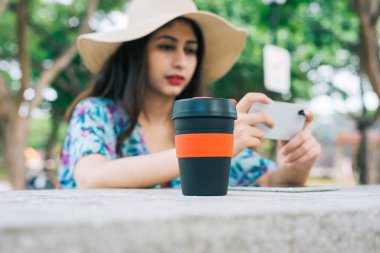 Açık hava parkında elinde cep telefonu olan modern bir Hintli kadın. Masada çevre dostu yeniden kullanılabilir bir fincan ve dokunmatik ekran tableti var..