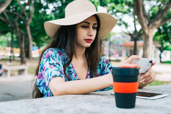 Reusable cup in foreground. Background blur Indian female in outdoor park holding mobile phone device. She could be seen working out of office, video call with families & friends, or watching video.