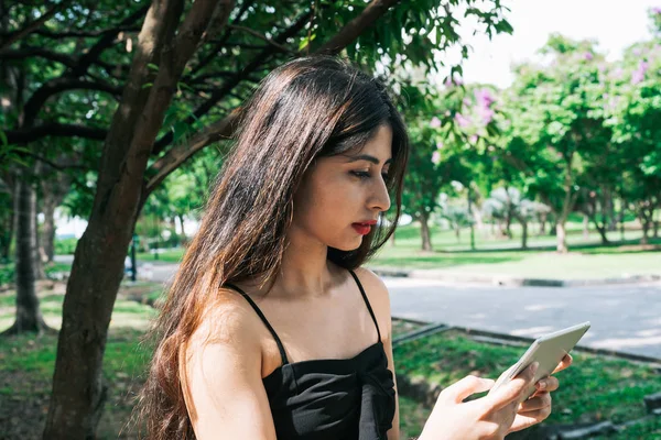 One long hair Indian girl holding and using mobile touch screen device tablet at outdoor park