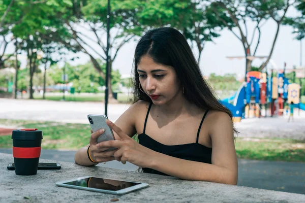 Um cabelo longo indiana fêmea segurando telefone celular no parque ao ar livre. Na mesa está um tablet touch screen e um copo reutilizável eco-friendly . Imagem De Stock