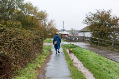Islak ve yağmurlu bir günde orta yaşlı bir kadın nehir yolunda küçük köpeğini dolaştırıyor.