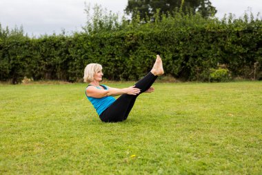 Çimenli bir parkta çıplak ayak yoga yapan orta yaşlı bir kadın. O parlak mavi bir yelek ve siyah tayt giyiyor. Yoganın stili Hatha Yoga.