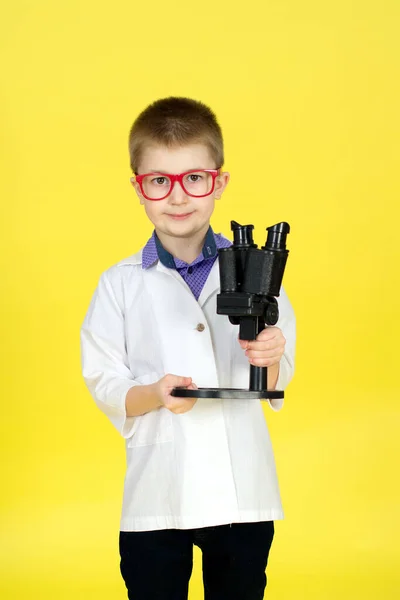 Pojke med glasögon, medicinsk klänning och mikroskop på gul bakgrund. Liten forskare gör upptäckter, utför experiment. Letar efter ett botemedel, ett vaccin mot viruset. Begreppet framtid — Stockfoto