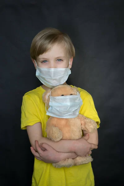 Criança em uma máscara médica com um urso de brinquedo em seus braços em um fundo preto. O jogo do médico. O conceito de proteção contra vírus e doenças. Parem o coronovírus. Covid-19 — Fotografia de Stock