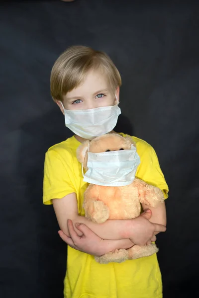 Niño con una máscara médica con un oso de juguete en los brazos sobre un fondo negro. El juego del doctor. El concepto de protección contra virus y enfermedades. Detén el coronovirus. Covid-19 — Foto de Stock