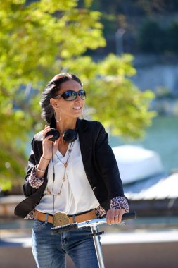 Güneş gözlüklü, kulaklıklı, motosikletli genç ve güzel bir kız yazın yeşil nehir ve ağaçlarla dolu bir sokakta.