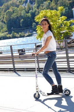 Genç ve güzel bir kız yazın yeşil nehir geçmişiyle sokakta scooter kullanıyor.