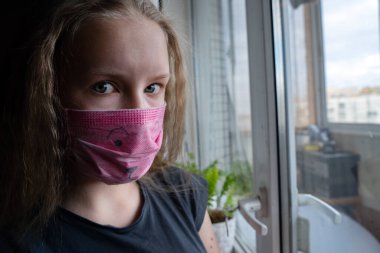 Tıbbi maske takan genç kız portresi Covid-19 karantina sırasında pencerenin yanındaki kameraya bakın.