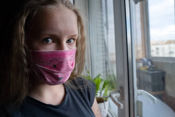 Tıbbi maske takan genç kız portresi Covid-19 karantina sırasında pencerenin yanındaki kameraya bakın. — Stok fotoğraf
