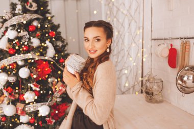 Güzel bir kız beyaz bir fincandan kahve ya da çay içiyor. Noel sabahı havası, genç bayana yakın noel ağacı.