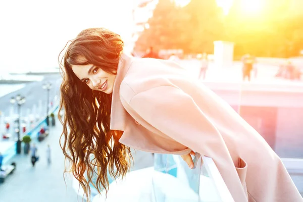 Beautiful young girl with dark hair stands near the sea in a beige coat and smiles. — Stock Photo, Image