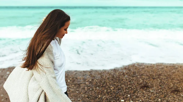 Una hermosa joven camina a lo largo de la orilla del mar, una tormenta, el pelo vuela aparte, un cárdigan gris, una figura deportiva en zapatillas de deporte disfruta, otoño o invierno — Foto de Stock