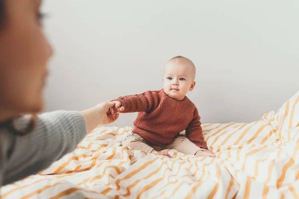 Linda menina na cama em um acolhedor sorriso camisola marrom. Conceito de maternidade e infância. Mãe segurar pequena mão . — Fotografia de Stock
