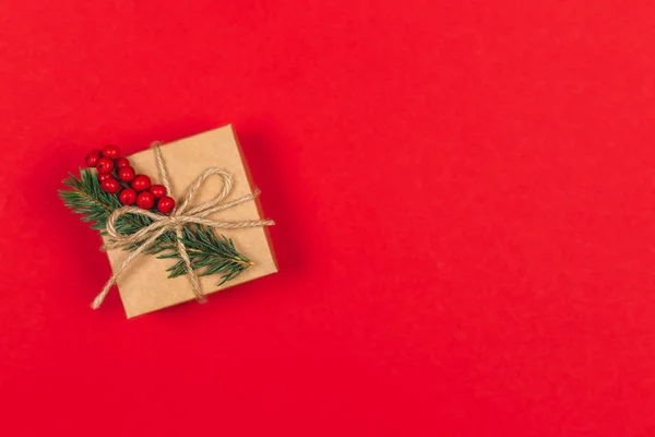 Caja de regalo artesanal con rama de abeto y bayas rojas sobre fondo rojo. Concepto ecológico de vacaciones . — Foto de Stock