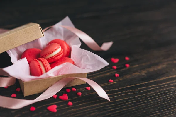 Macarons franceses vermelhos em uma caixa, decorados com corações doces e fita em mesa de madeira escura. Conceito para o dia dos namorados . — Fotografia de Stock